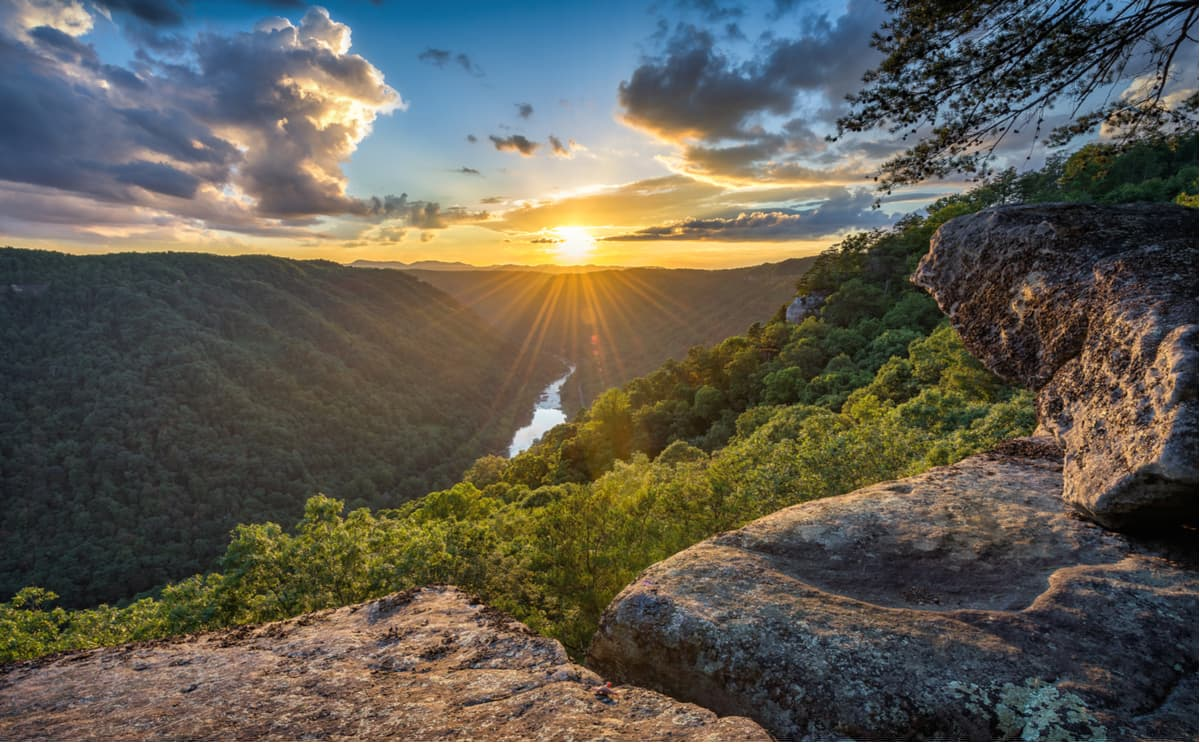 Sunset over an Appalachian river gorge — sunrays breaking through clouds above forested ridgelines, viewed from a rocky overlook.