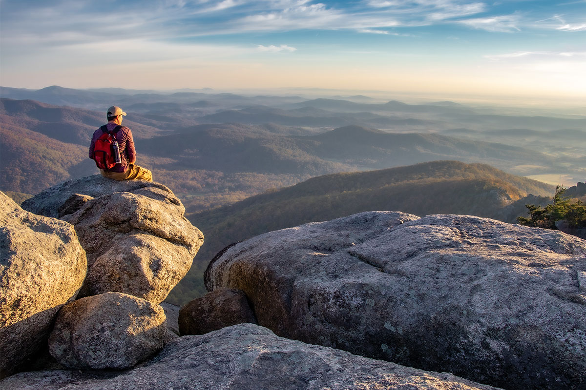 A hiker resting on the granite summit of Old Rag Mountain in Shenandoah National Park, Virginia, looking out over layered Blue Ridge ridgelines at golden hour.